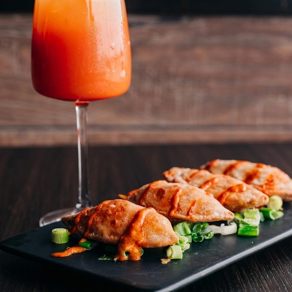 Four empanadas with orange sauce and green onions on a black plate, next to an orange drink.