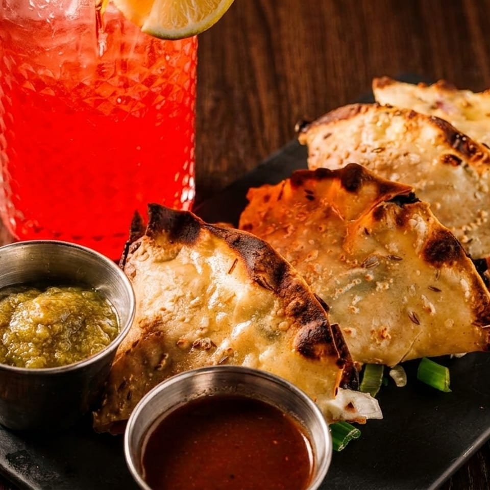Empanadas with green and red sauces, alongside a red cocktail with a lime wedge.