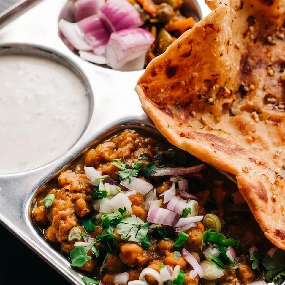 A metal tray holds chana masala, naan with butter, raita, pickled onions, and mixed vegetables.